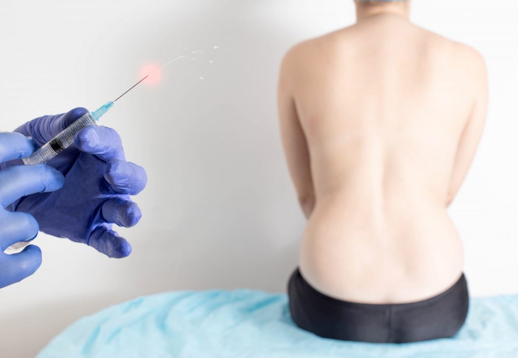 A person is seated with their back to the camera, revealing their bare upper body, while a healthcare professional in gloves prepares a syringe with a visible drop at the needle tip for facet joint injections,