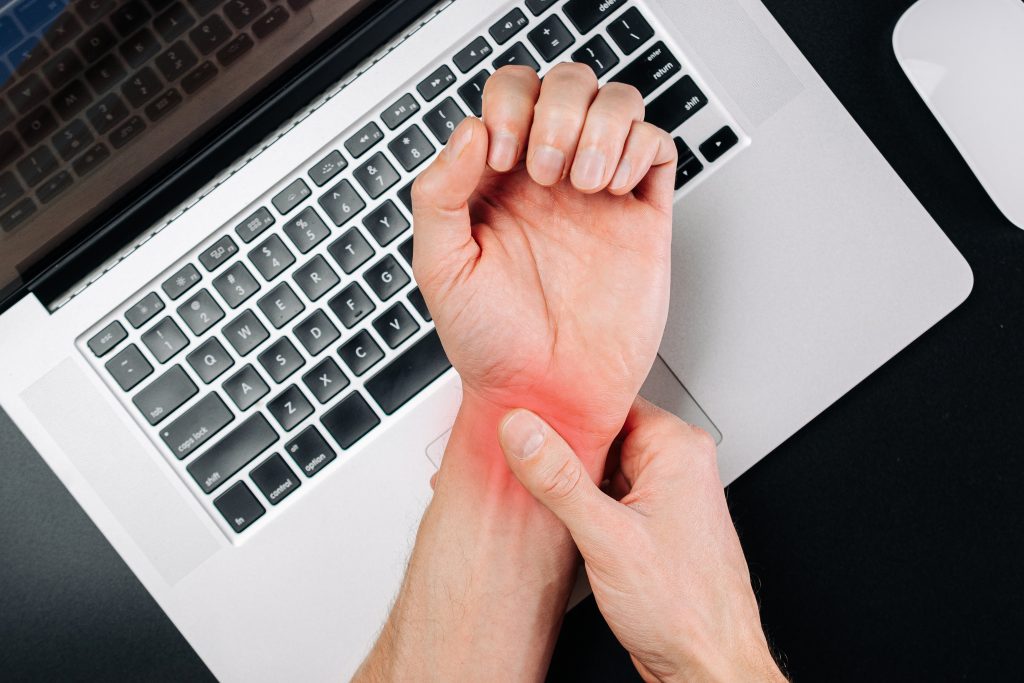 A person experiencing wrist pain while working on a laptop, possibly indicative of repetitive strain injury (RSI) or needing carpal tunnel surgery.