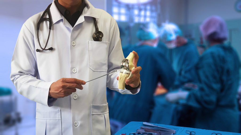 A male surgeon in a white coat with a stethoscope holding a model of a knee joint, with a blurry background of a surgical team in an operating room.