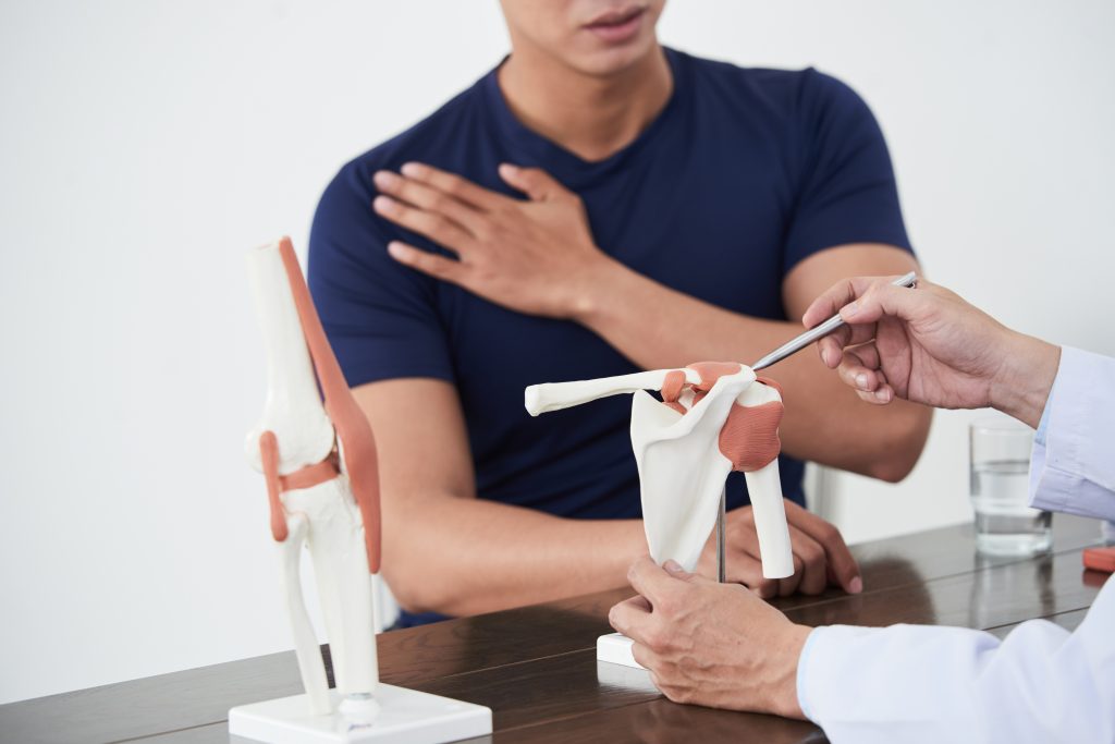 A doctor explains a shoulder joint model to a male patient, who listens attentively in an examination room.