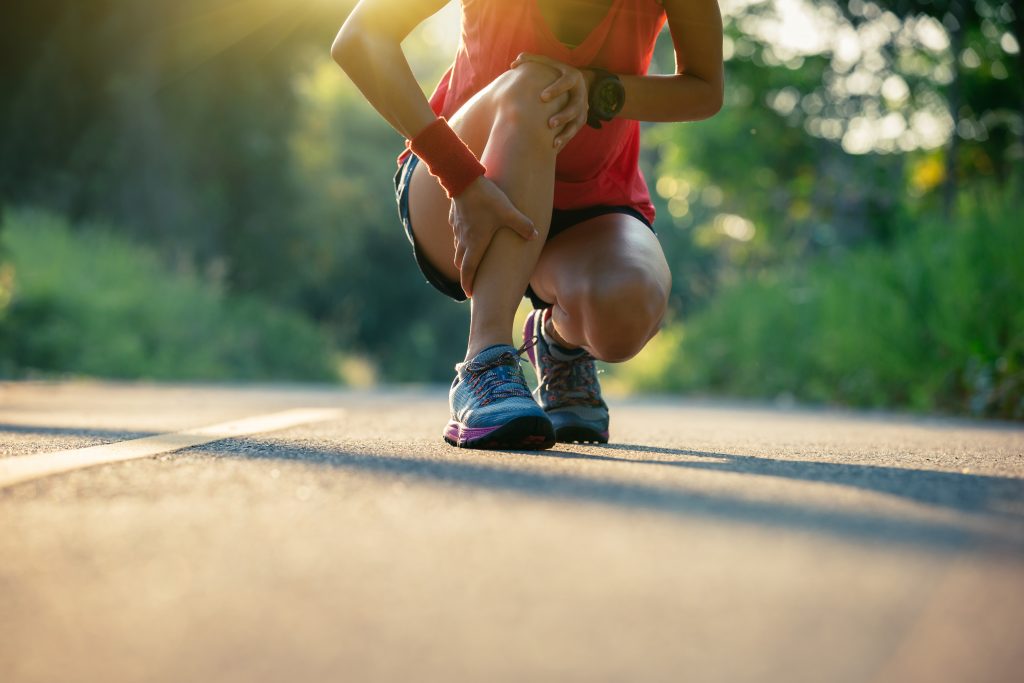 A person in sportswear crouches on an outdoor trail, holding their lower leg as if in pain, possibly from a sports injury. They are wearing a red top, wristband, and watch. The scene is set during a sunny day with trees visible in the background.