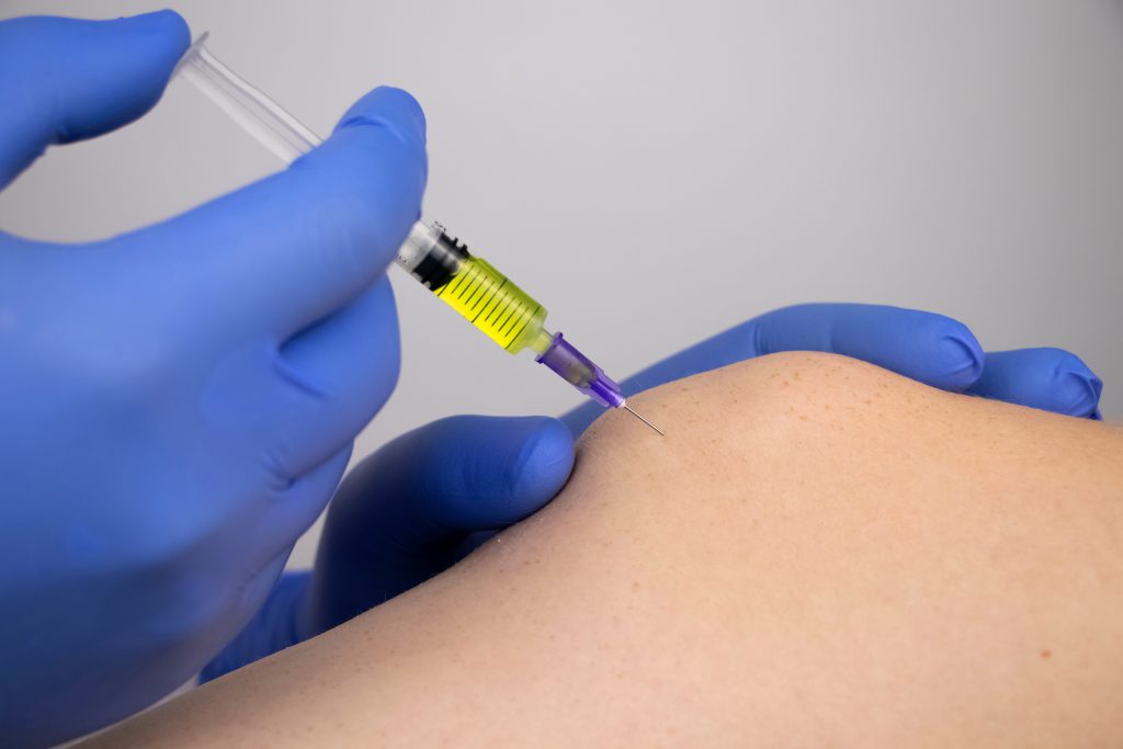 Close-up of gloved hands administering a vaccine injection to a person's upper arm. The syringe, partially filled with yellow liquid, suggests immediate relief often seen in corticosteroid injections. The needle is inserted into the skin, with only the person's shoulder and upper arm visible.