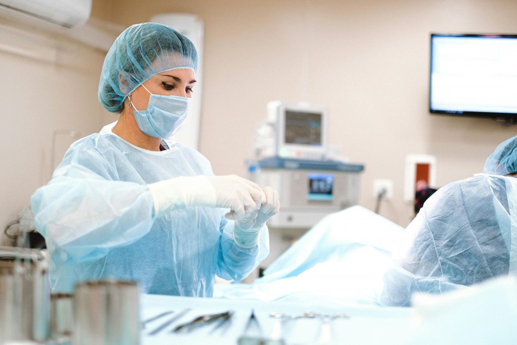 A medical professional wearing a blue surgical gown, mask, and cap prepares instruments in an operating room. Another healthcare worker, involved in a cervical fusion recovery case, is partially visible in the background next to medical equipment and a monitor displaying information.