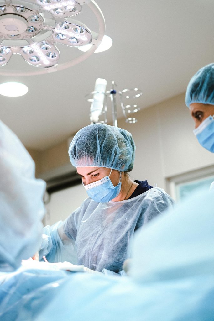 Medical professionals wearing blue surgical gowns, masks, and caps performing knee surgery under bright operating room lights. An overhead surgical light illuminates the scene. Their focused expressions are partially visible through their masks, indicating the essential must-haves for a successful recovery.