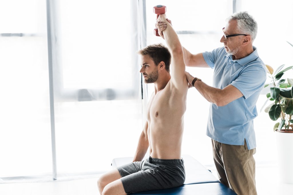 chiropractor standing near man exercising with dumbbell in clinic