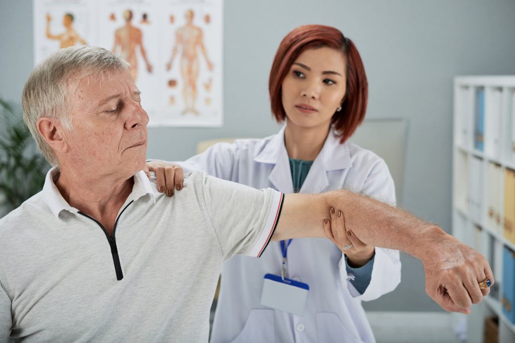 An older man with gray hair is having his arm examined and moved by a healthcare professional with short red hair, who is wearing a white lab coat and a name badge. They are in a medical office with anatomical posters on the wall in the background, discussing types of shoulder operations.